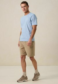Lichtblauw katoenen t-shirt met korte mouwen, gecombineerd met beige shorts. Model draagt grijze sneakers en opgerolde sokken, staand voor een neutrale achtergrond.