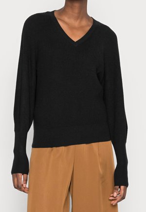 Schwarzer Strick-V-Ausschnitt-Pullover mit langen, schmal zulaufenden Ärmeln und geripptenDetails. Kombiniert mit lockeren, tanfarbenen Hosen, die eine entspannte Passform zeigen.