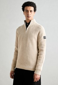 Suéter de ribete beige con cuello de media cremallera, con un tejido texturizado. Incluye una etiqueta negra en la manga izquierda.