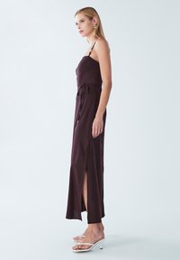 Abito lungo senza maniche di colore viola scuro con spacchi laterali, dettaglio con cintura annodata in vita e spalline sottili. Indossato con sandali bianchi con tacco alto.