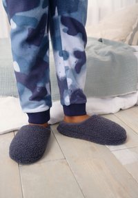 Lila Fleecepantoffeln mit einer weichen, strukturierten Oberfläche und abgerundeter Form, kombiniert mit blau gemusterten Schlafanzughosen und einem neutralen Bettwäschehintergrund.