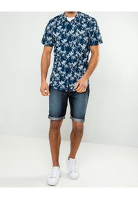 Camicia a maniche corte, blu navy, con motivo di palme bianche; abbinata a pantaloni shorts di jeans arrotolati e sneakers bianche.