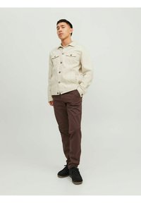Beige denim jacket with two chest pockets and button closure, paired with brown trousers and dark shoes. Simple design, smooth texture.