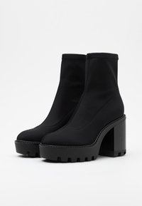Botas negras hasta el tobillo con una plataforma gruesa y un alto tacón cuadrado. Hechas de un material sintético suave con un diseño ajustado y costuras mínimas.