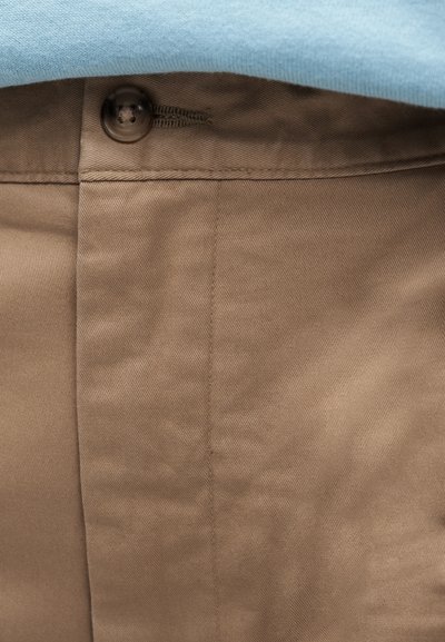 Κοντινό πλάνο καφέ παντελονιού από ύφασμα με σκούρο κουμπί και μέρος ενός ανοιχτό μπλε πουκαμίσου ορατό στη μέση.