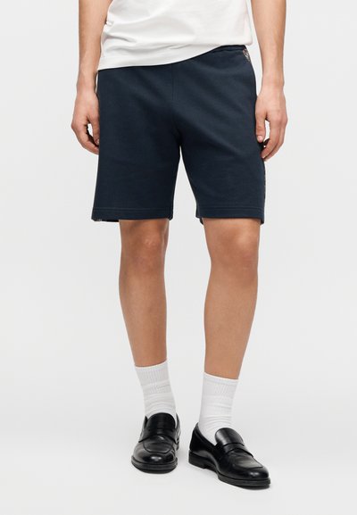 Σκούρα μπλε αθλητικά σορτς με ελαστική μέση, διαθέτουν πλαϊνές τσέπες και διακριτική υφή. Φορεμένα με λευκές κάλτσες και μαύρα μοκασίνια.