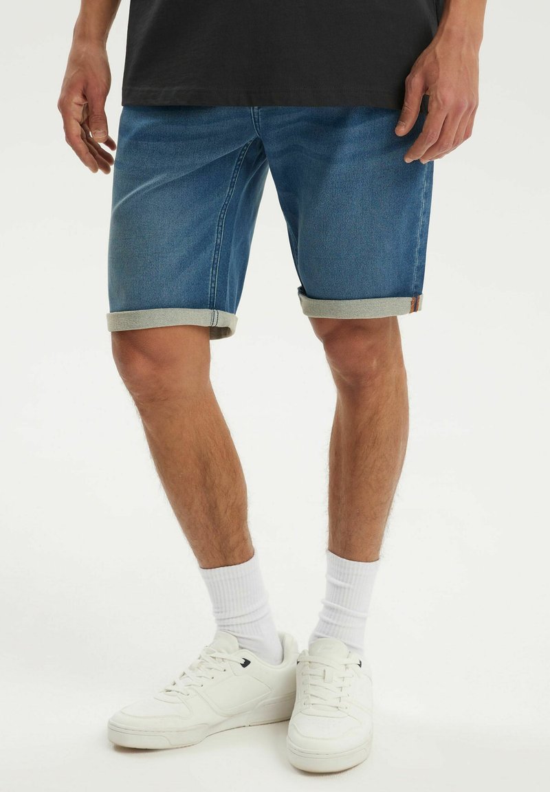 Mężczyzna w podwiniętych niebieskich jeansowych krótkich spodenkach, białych skarpetkach i białych trampkach, stojący na jednolitym tle.