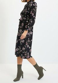 Kobieta w czarnej midi sukience w różowy kwiatowy wzór oraz oliwkowych wysokich botkach-skarpetkach, idąca na tle jednolitego białego tła.