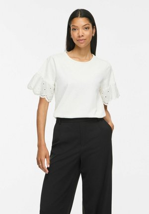 Femme aux longs cheveux foncés portant un chemisier blanc à manches courtes avec des manches en dentelle à œillets et un pantalon noir taille haute à jambes larges.