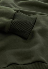 Mørkegrønt sweatshirtstof med ribbet manchet detaljer vist i et nærbillede.