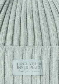 Lichtgroene geribbelde gebreide beanie met een textuurfinish en een rechthoekig stoffen label met de tekst "VIND JE INNERLIJKE VREDIGHEID" en "vertrouw op je ontwerp."
