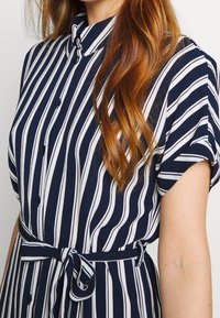Abito a righe navy e bianche con bottoni, maniche corte, colletto e cintura con nodo in vita. Tessuto liscio con un motivo testurizzato.