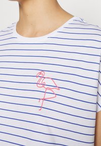 Camiseta blanca con rayas horizontales azules y un diseño bordado de un flamenco rosa en el área del pecho izquierdo. Corte regular y mangas cortas.