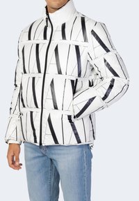 Giacca a vento bianca con strisce verticali nere, chiusura con zip, collo alto e due tasche laterali. Indossata sopra un denim blu.