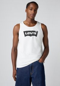 Bjela pamučna majica bez rukava s crnim Levi's logotipom u središtu. Uparena s tamnoplavim trapericama. Jednostavan, ležeran dizajn.