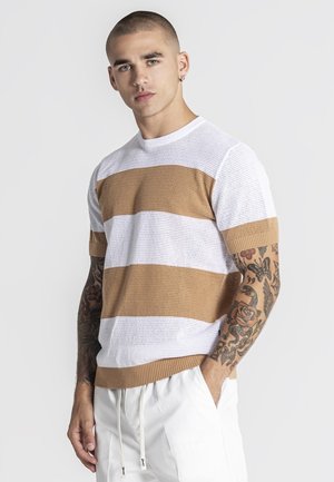 Mladý muž s tetovaniami, oblečený v bielom a béžovo pruhovanom pletenom tričku a bielych nohaviciach so šnúrkou, stojaci s jednou rukou v vrecku.