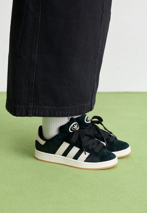 Baskets Adidas noires avec des bandes blanches et de gros lacets noirs portées avec des chaussettes blanches et un pantalon noir sur un sol vert clair.