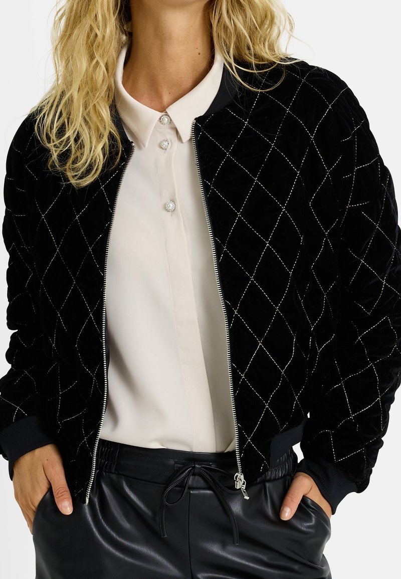 Femme portant une veste matelassée noire zippée sur un chemisier blanc boutonné, avec un pantalon en cuir noir et les mains dans les poches.