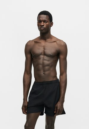 Homme athlétique debout devant un fond uni, portant un short de sport noir, torse nu, avec une expression faciale neutre.