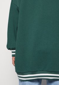 Sweat-shirt vert oversize avec poignets côtelés, présentant des rayures blanches et vert foncé à l'ourlet et aux manches. Tissu doux et texturé.