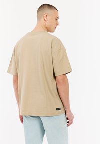 Beige t-shirt met korte mouwen van katoen, met een relaxte pasvorm en een label aan de onderzijde links. G gedragen met lichtblauwe denim jeans.