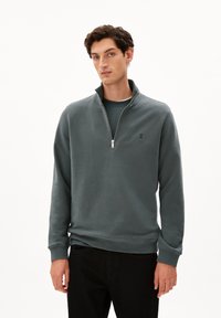 Grå zip-up sweatshirt med hög krage, med en mjuk textur och en subtil logotyp på bröstet, i kombination med mörka byxor.