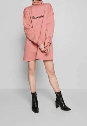 Person iført en lyserød oversized sweatshirtkjole med "Expensive" tekst og sorte ankelstøvler, stående mod en grå baggrund.