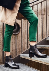 Bottes cheville en cuir noir à finition lisse, avec des panneaux latéraux élastiques et des talons carrés, assorties à un pantalon vert retroussé sur un escalier en métal.