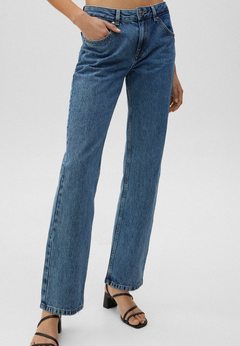 Jean denim bleu droit porté avec des sandales à talons noires à brides, une main dans la poche, sur un fond clair uni.