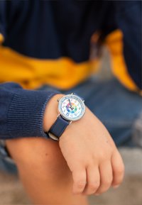 Montre colorée pour enfants avec un cadran rond, un cadran blanc présentant des chiffres et des couleurs, et un bracelet en cuir bleu. Main reposant sur un genou.