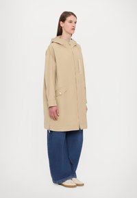 Giacca impermeabile beige con cappuccio, zip frontale e tasca a toppa. Abbinate a pantaloni in denim blu a gamba larga e sneakers di colore chiaro.