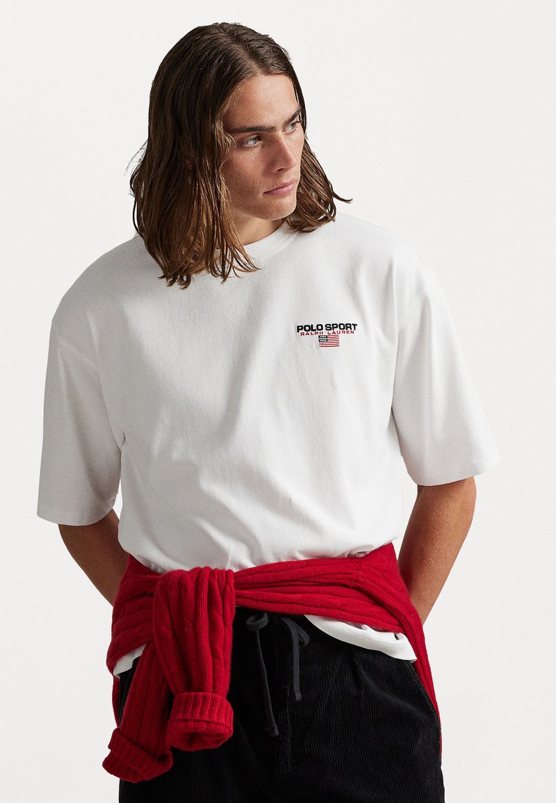 Joven con cabello hasta los hombros que lleva una camiseta blanca de Polo Sport Ralph Lauren y un suéter rojo atado alrededor de la cintura.