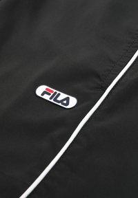 Primer plano de tela negra con ribete blanco y un parche con el logo de FILA en blanco, rojo y azul marino.