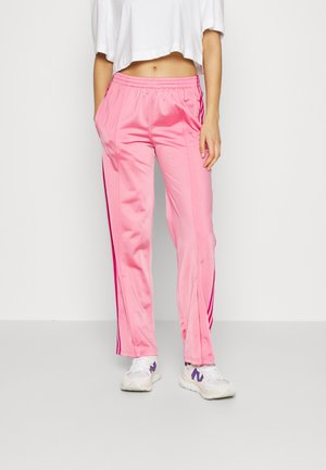 Femme portant un pantalon de survêtement rose brillant avec taille élastique et bandes latérales, un t-shirt blanc court, et des baskets blanches avec des accents violets.