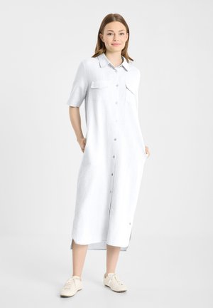 Vrouw staand, draagt een witte midi-jurk met knopen, korte mouwen, voorzakken en beige sneakers tegen een effen achtergrond.