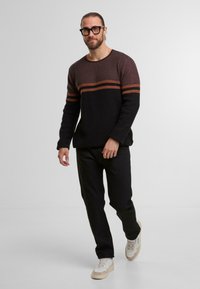 Czarny dzianinowy sweter z bordowymi i pomarańczowymi poziomymi paskami, zestawiony z czarnymi spodniami i jasnymi sneakersami. Model stoi w neutralnym otoczeniu.