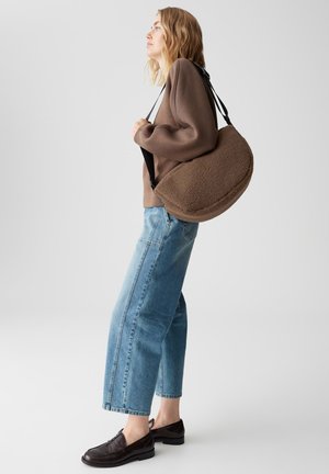 Donna con un maglione marrone e jeans a gamba larga porta una grande borsa a tracolla marrone texturizzata con tracolla nera.