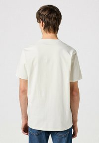 Camiseta de manga corta en color crema, hecha de algodón, con cuello redondo y un corte relajado, combinada con jeans de mezclilla azul oscuro.