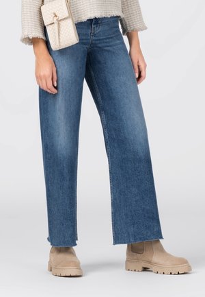 Femme portant un jean bleu à jambes larges, des bottes en daim beige, un pull texturé crème et portant un petit sac bandoulière tressé beige.