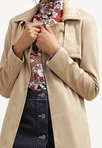 Beige mockaskinnjacka med spetsig krage, knäppning fram och två fickor. Bärs över en mönstrad blus med hög hals och jeanninskjorta.