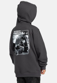 Grijze hoodie met een groot zwart-wit grafisch ontwerp op de achterkant, inclusief tekst en een afbeelding van een skateboarder.