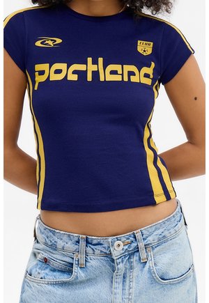 Frau trägt ein enges blaues Crop-Top mit gelben Streifen und dem Text "portland", kombiniert mit hellblauen hoch taillierten Jeans.