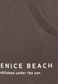 Brown fabric tank top featuring a ribbed texture. White printed text reads "VENICE BEACH established under the sun."