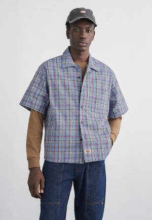 Mężczyzna w szaro-czerwonej kratowanej koszuli z krótkim rękawem na beżowej koszuli z długim rękawem, ciemnoniebieskich jeansach i czapce z daszkiem.