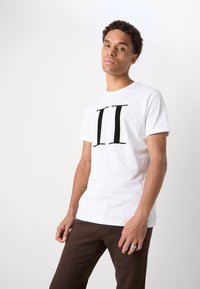Camiseta blanca de manga corta con un gran diseño gráfico negro "II" en el frente. Tejido suave y liso en combinación con pantalones marrones. Líneas simples y limpias.