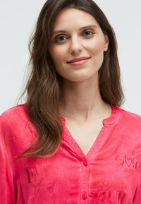 Frau mit langen braunen Haaren, die ein leuchtend pinkes button-up Hemd mit subtil bedrucktem Text und Designs trägt, lächelt sanft vor einem schlichten Hintergrund.