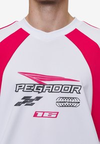 Biely športový dres s ružovými raglánovými rukávmi. Obsahuje logo "PEGADOR" a rôzne grafické potlače, vrátane čísel a dizajnu pneumatík.