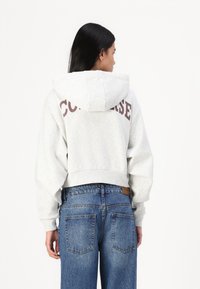 Light gray cropped hoodie with a large maroon "Converse" logo on the back, paired with high-waisted blue jeans featuring patch pockets.