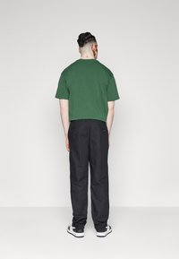 Green cotton short-sleeve t-shirt, black loose-fit trousers, and white sneakers, viewed from the back against a plain background.