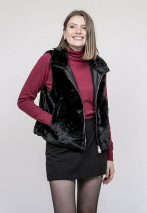 Femme souriante portant un gilet en fausse fourrure noire par-dessus un col roulé rouge, une jupe noire et des collants noirs transparents, debout devant un fond uni.
